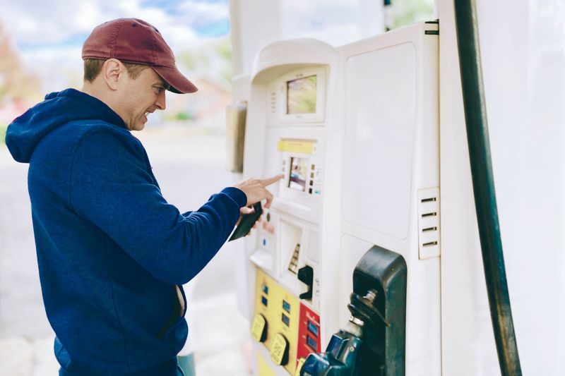 Man in blue hoodie and maroon baseball cap selecting payment method at a gas pump. 