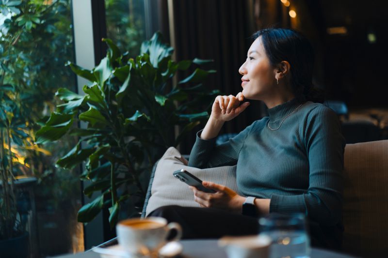 Confident young Asian woman with smartphone looking out through window while sitting in a cafe having coffee.