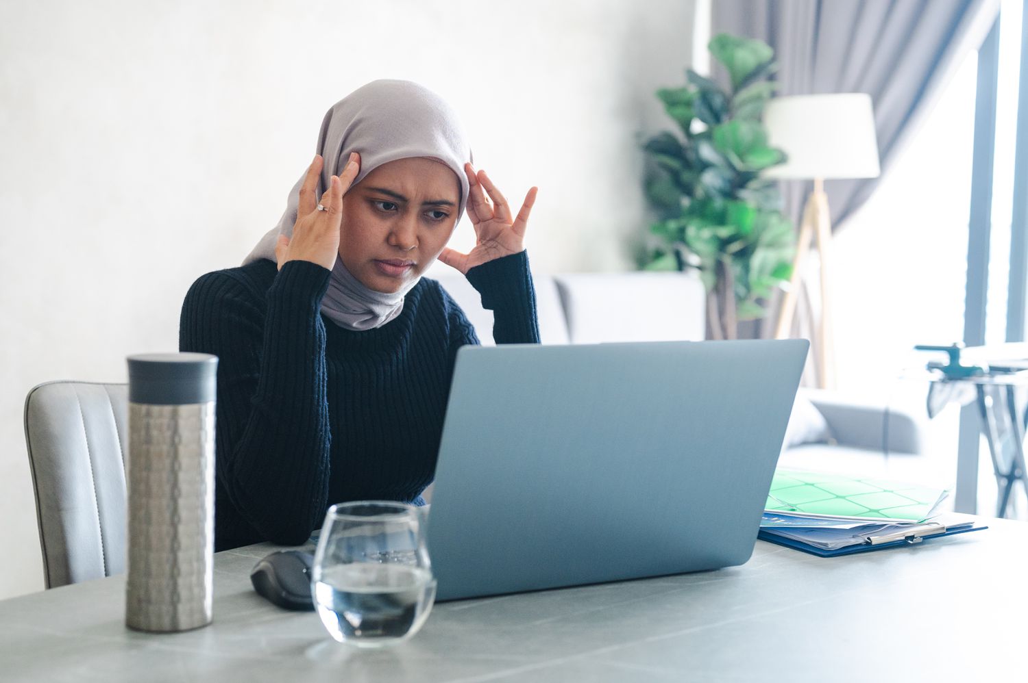 A young woman wearing a hijab frowns at her laptop screen. 