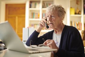 An older person on a phone while sitting in front of a laptop