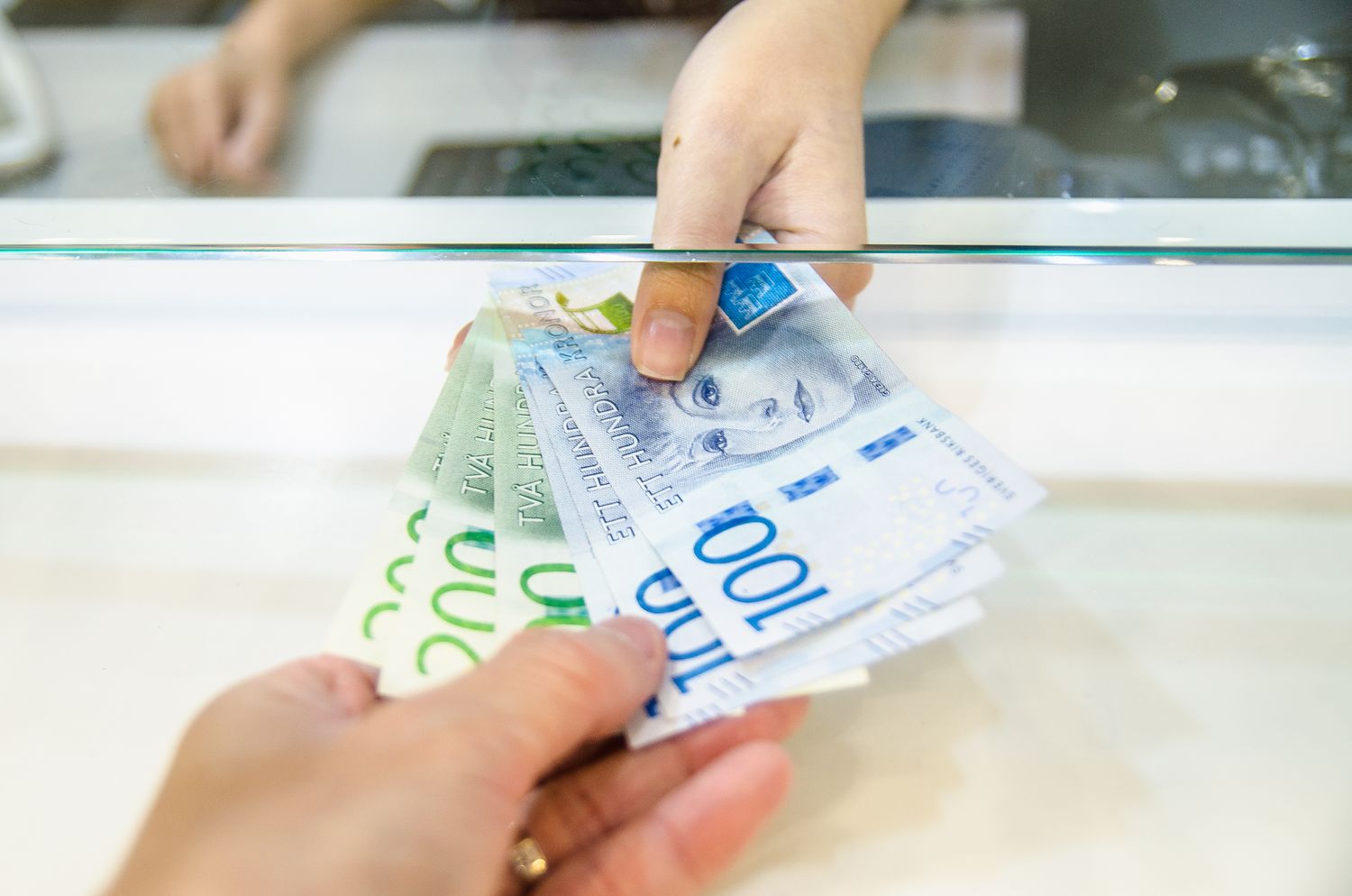 Two people exchange Swedish krona banknotes at a bank teller's window.