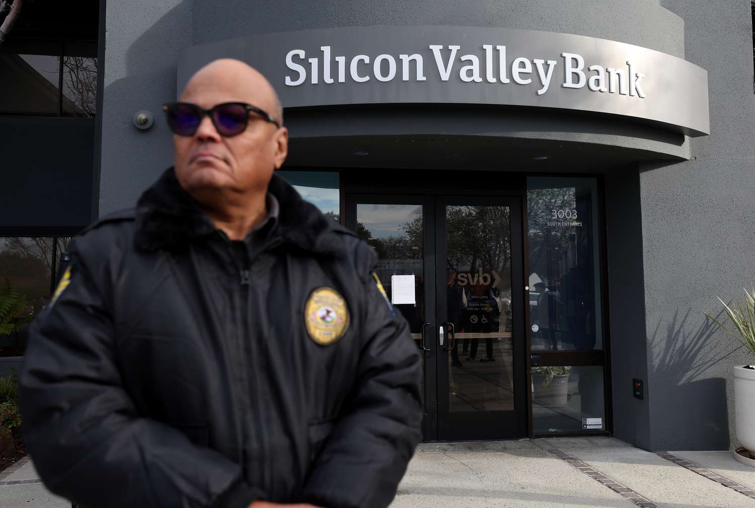 A security guard at Silicon Valley Bank monitors a line of people outside the office on March 13, 2023 in Santa Clara, California. Days after Silicon Valley Bank collapsed, customers are lining up to try and retrieve their funds from the failed bank. The Silicon Valley Bank failure is the second largest in U.S. history.