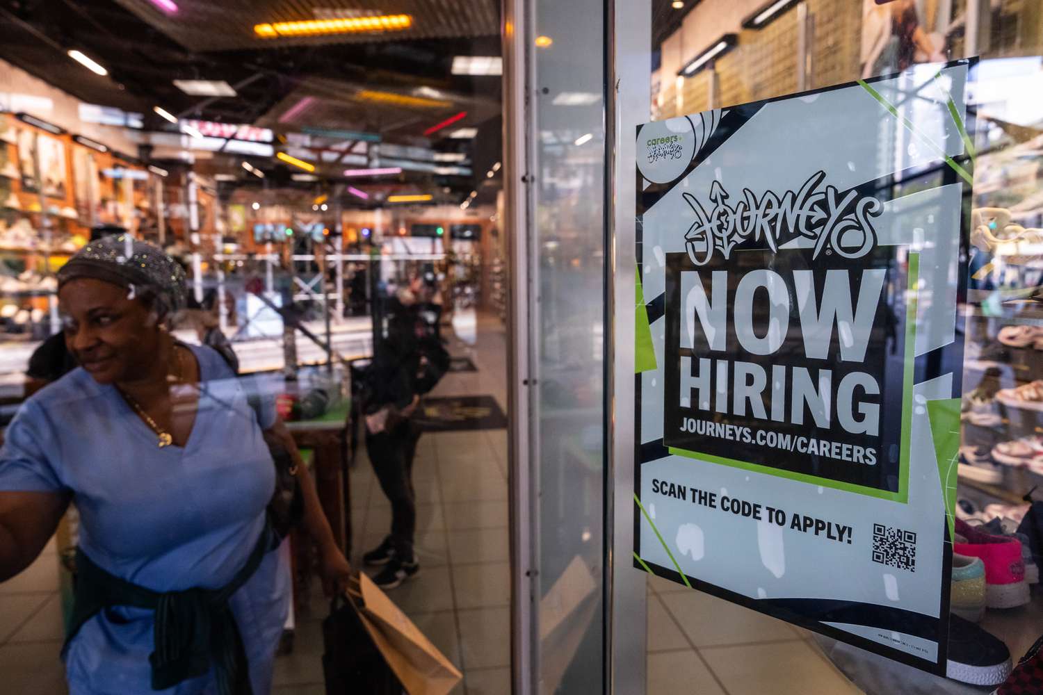 Journeys store entrance with a 'Now Hiring' sign, woman walking in the background, other store visitors visible