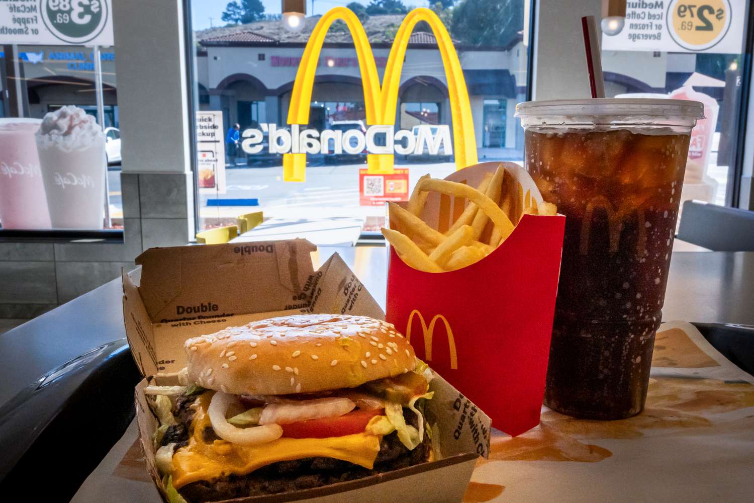A double quarter pounder with cheese, French fries, and a large drink sit on a table in front of a window with the McDonald's logo.