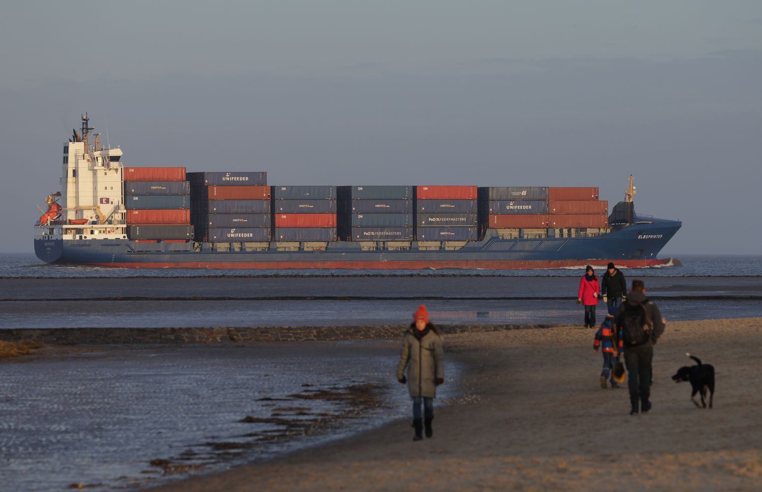 People stroll on the beach as a container ship passes by on February 05, 2023 in Cuxhaven, Germany. Germany's North Sea ports of Bremen, Bremerhaven and Hamburg are major shipping points for imports and exports.