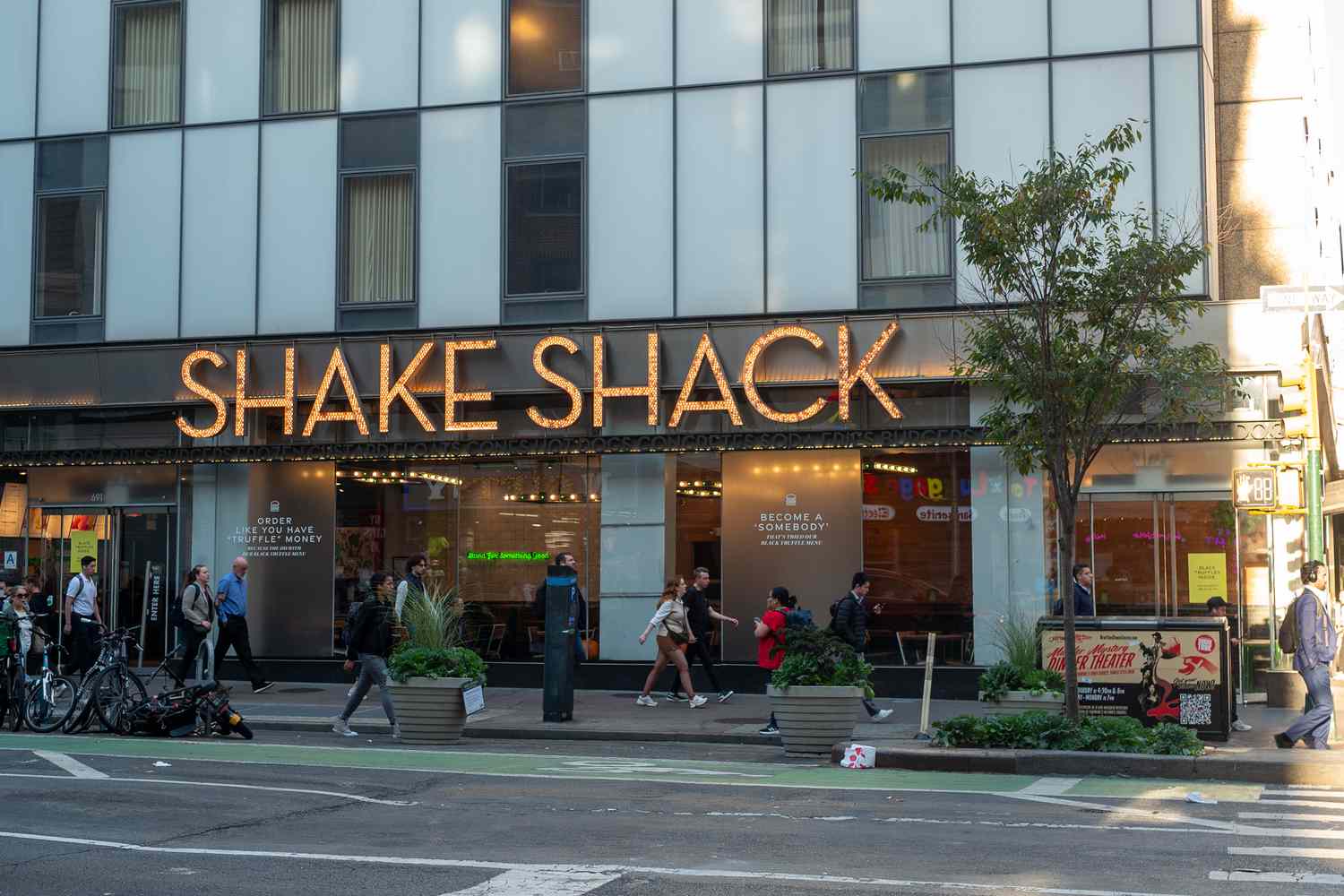 People walk on a sidewalk below the sign for a Shake Shack restaurant.