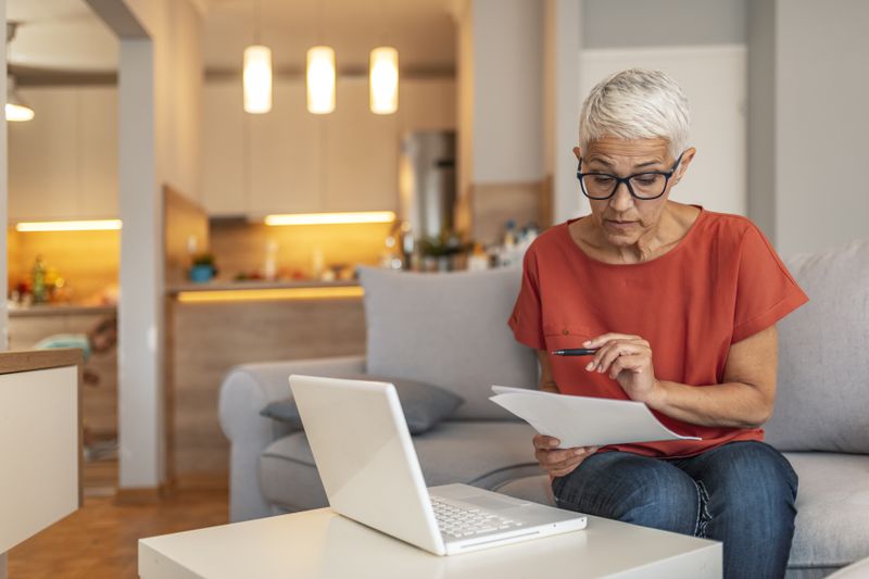 Mature woman in eyeglasses looking at her expenses using a laptop, pen, and paper.