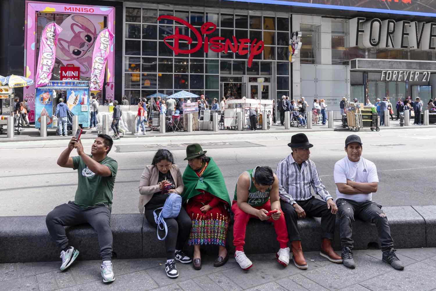 People sitting on a stone ledge in a city setting with a Disney store in the background, holding phones or in conversation