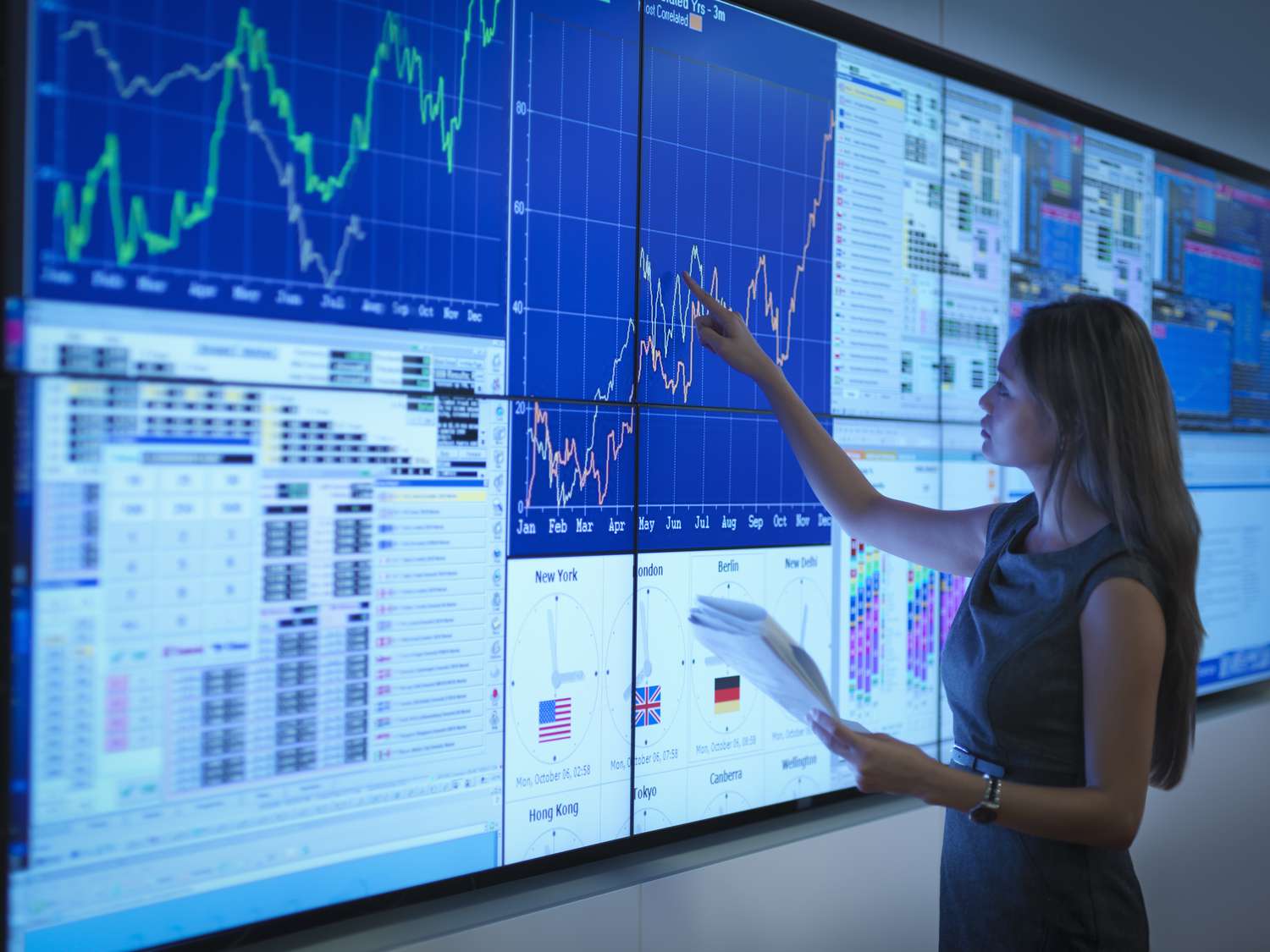 Woman standing in front of a wall of computer monitors, pointing to a chart. 