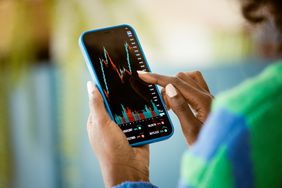 A close-up of a woman's hand, holding a cell phone and pointing to a stock chart on the screen. 