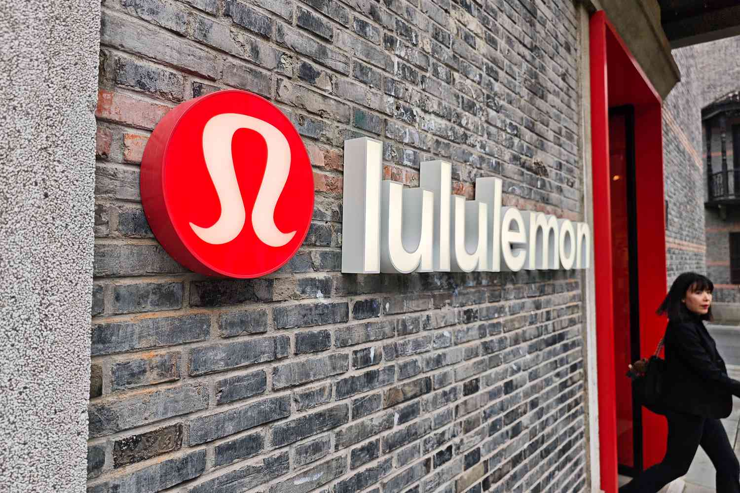 The red Lululemon logo is seen on the side of a store, with a person exiting the building.
