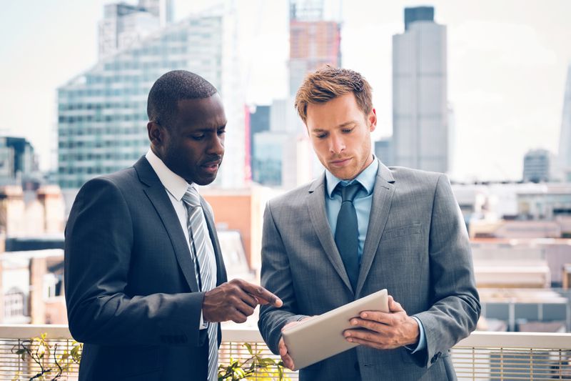 two men in suits looking at a tablet outside with a skyline in the background
