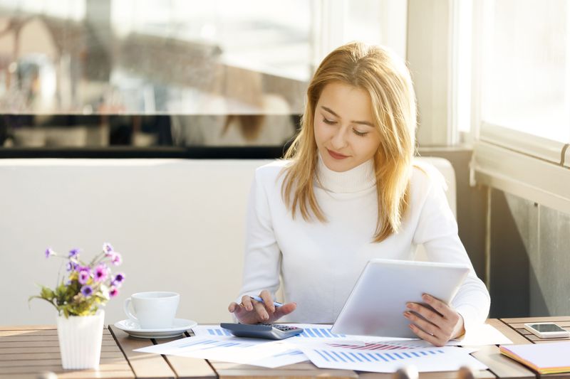 Businesswoman using calculator in sunny office space with printed out graphs on desk