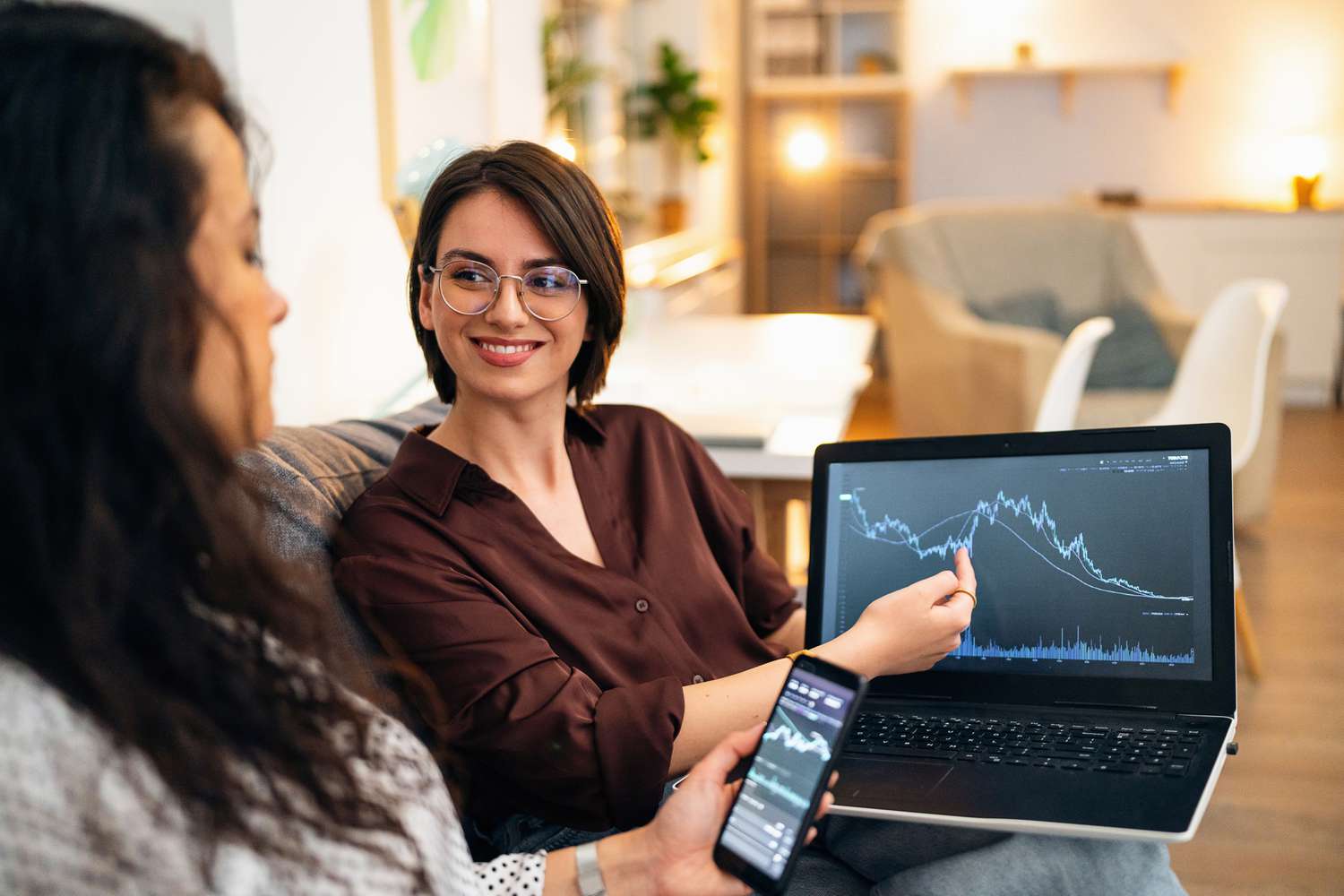 Two women sitting on a couch. One woman is showing the other a chart on her laptop.