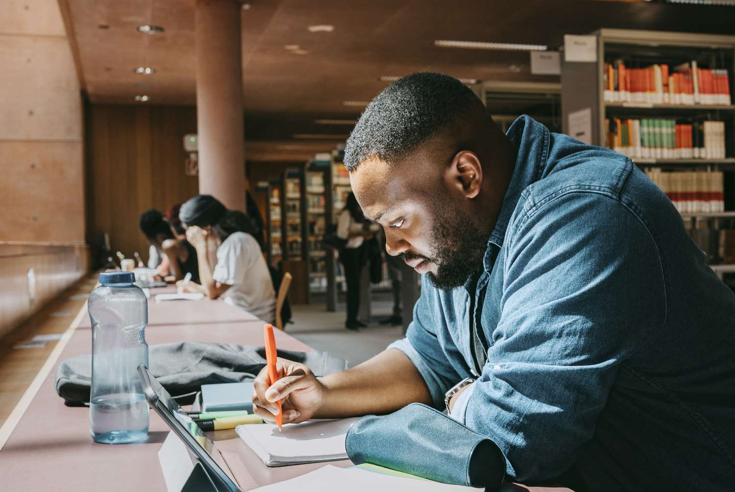 Student writing in notebook while sitting with tablet in college library