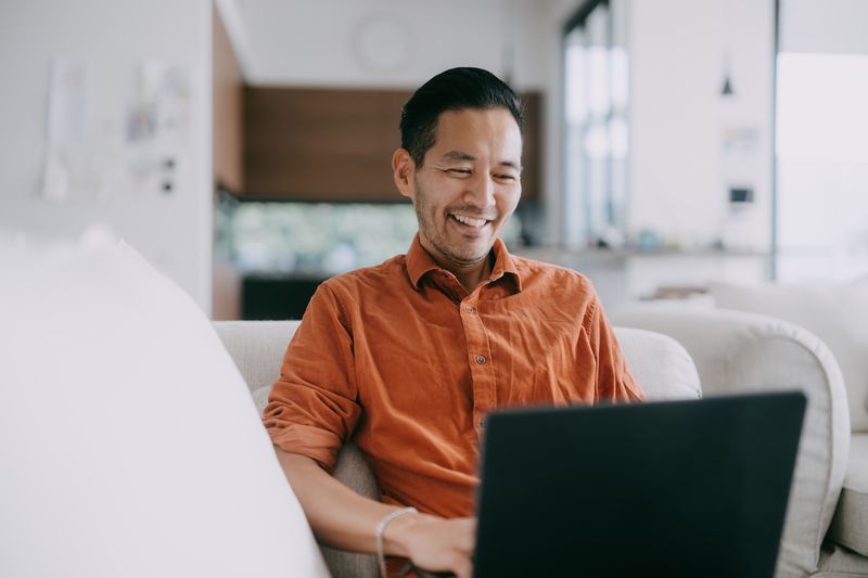 A happy man looks at his laptop while sitting on a couch.