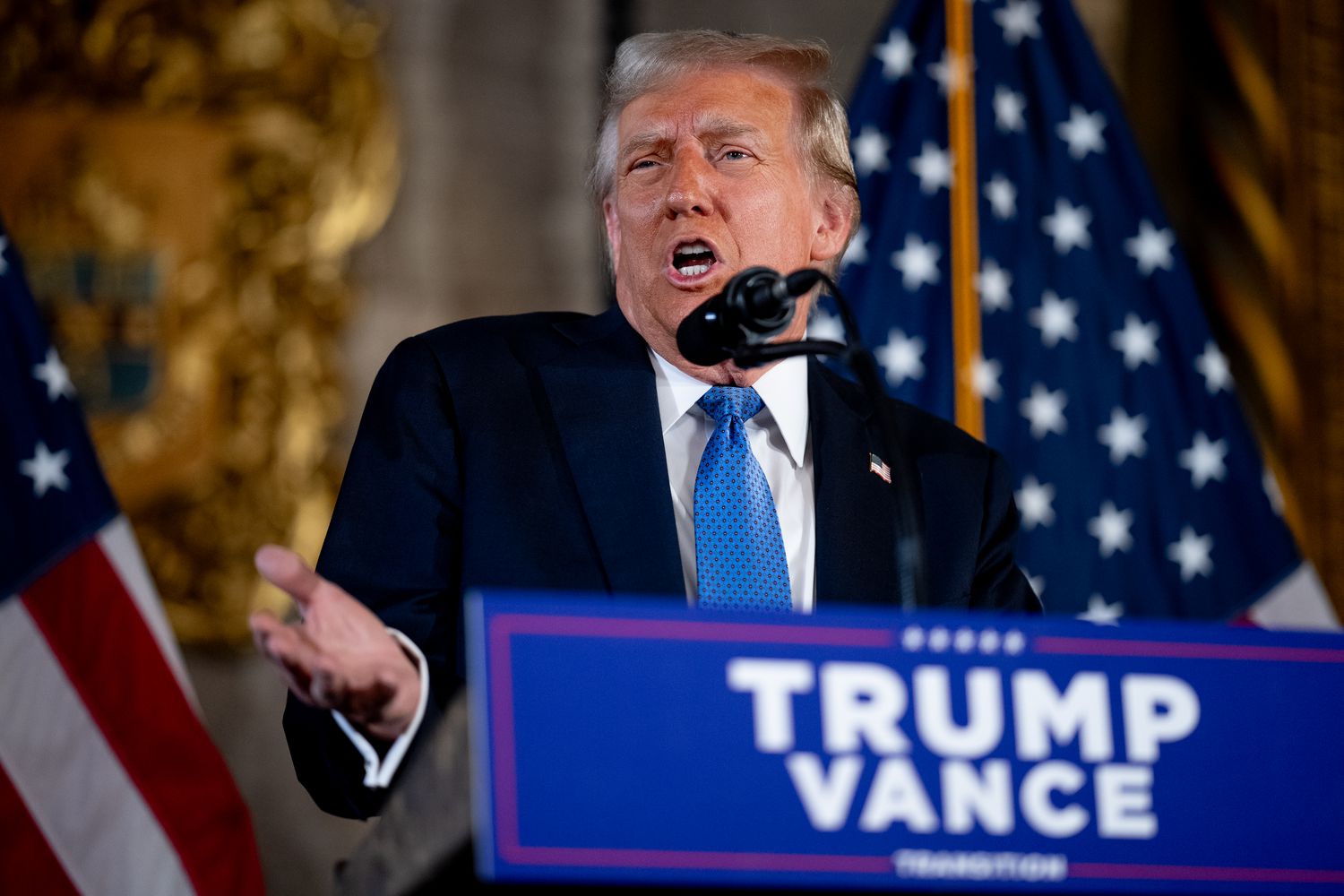 Donald Trump is seen standing at a Trump Vance podium in front of a microphone, with a pair of American flags in the background.