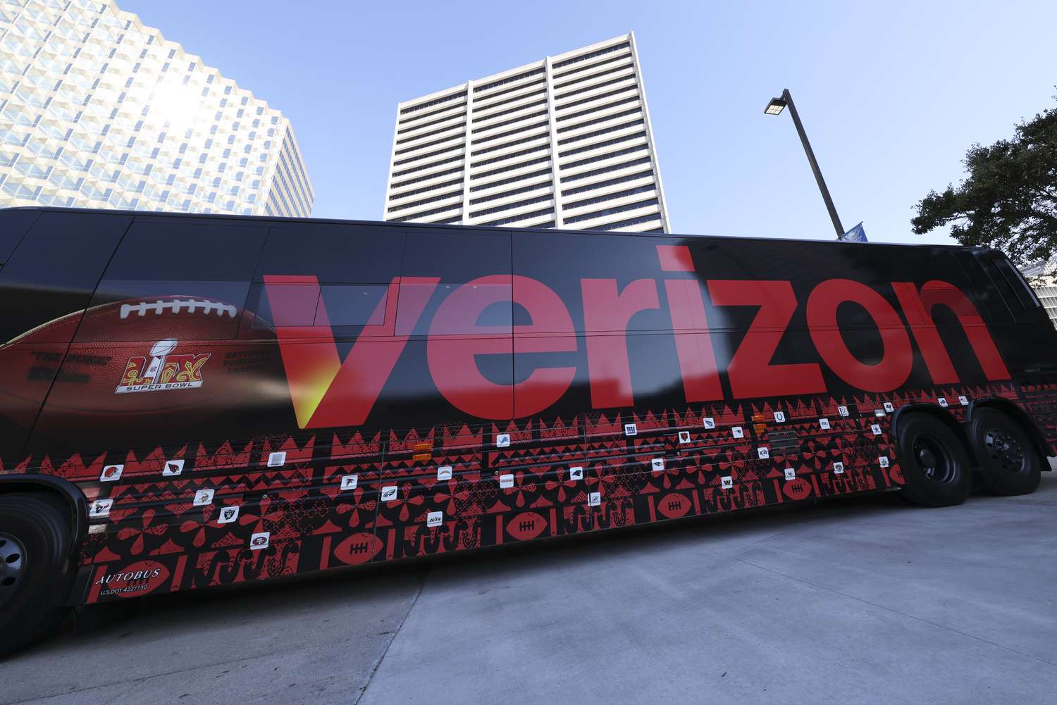 The Verizon logo is seen on the side of a bus with buildings and a blue sky in the backrgound.