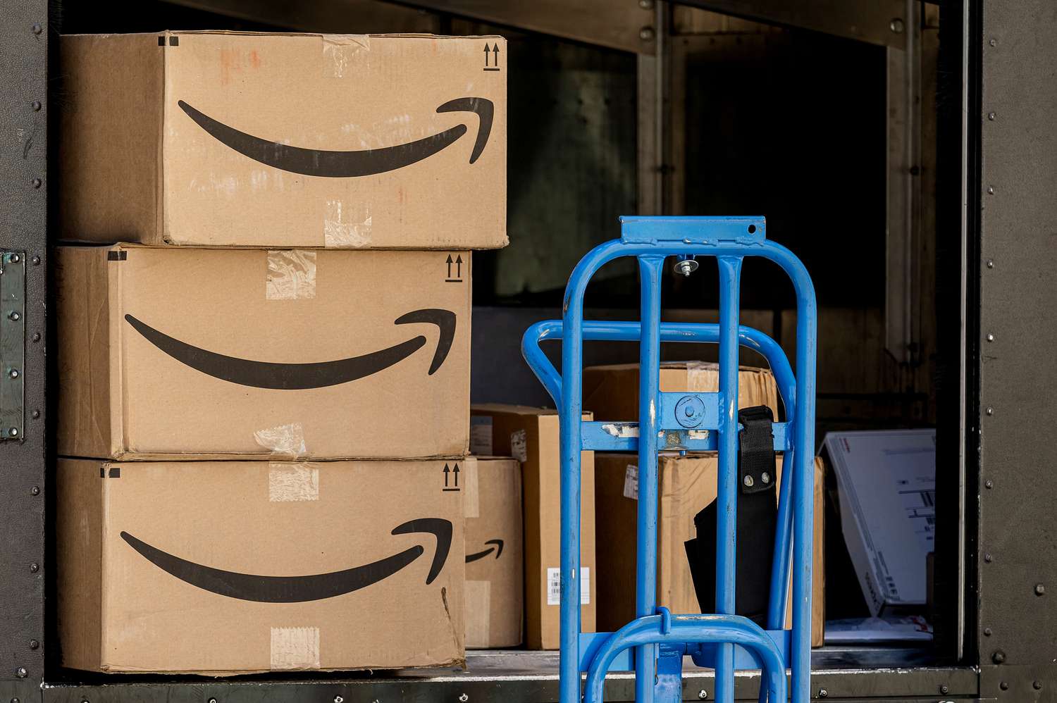 Amazon packages are seen inside a truck, with a blue dolly next to them.