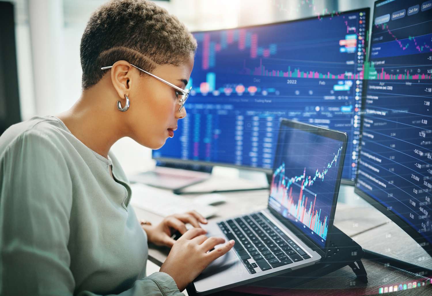 Female traders sitting at desk working on a laptop. Several monitors showing financial data are behind the laptop.