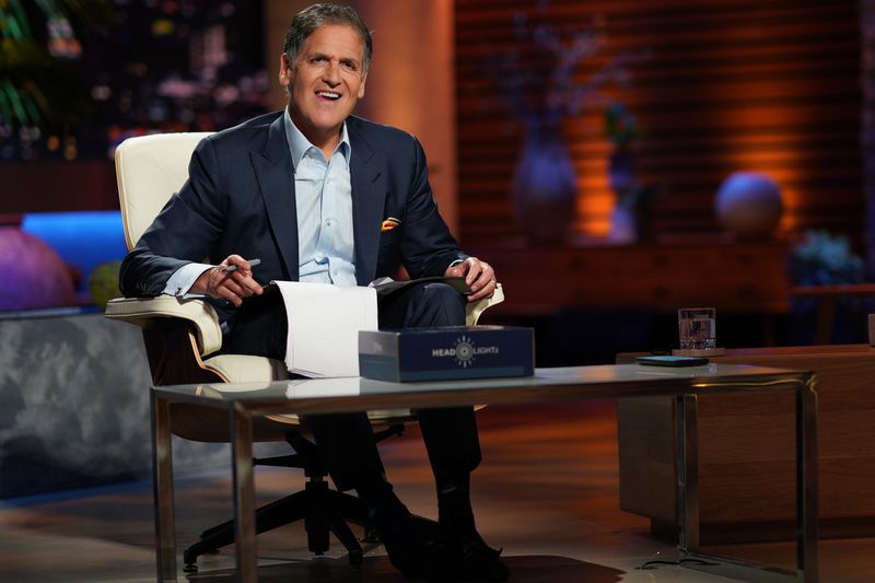 Mark Cuban sits in a chair with a notepad in his lap on the set of the TV show Shark Tank. 