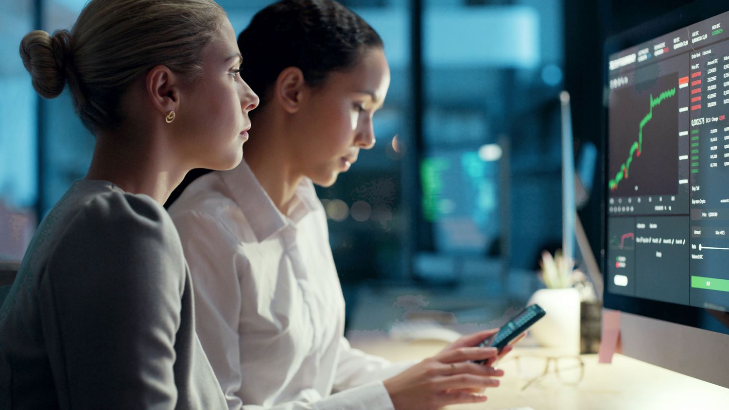 Two women looking at a smartphone and computer screen with financial market information on them.