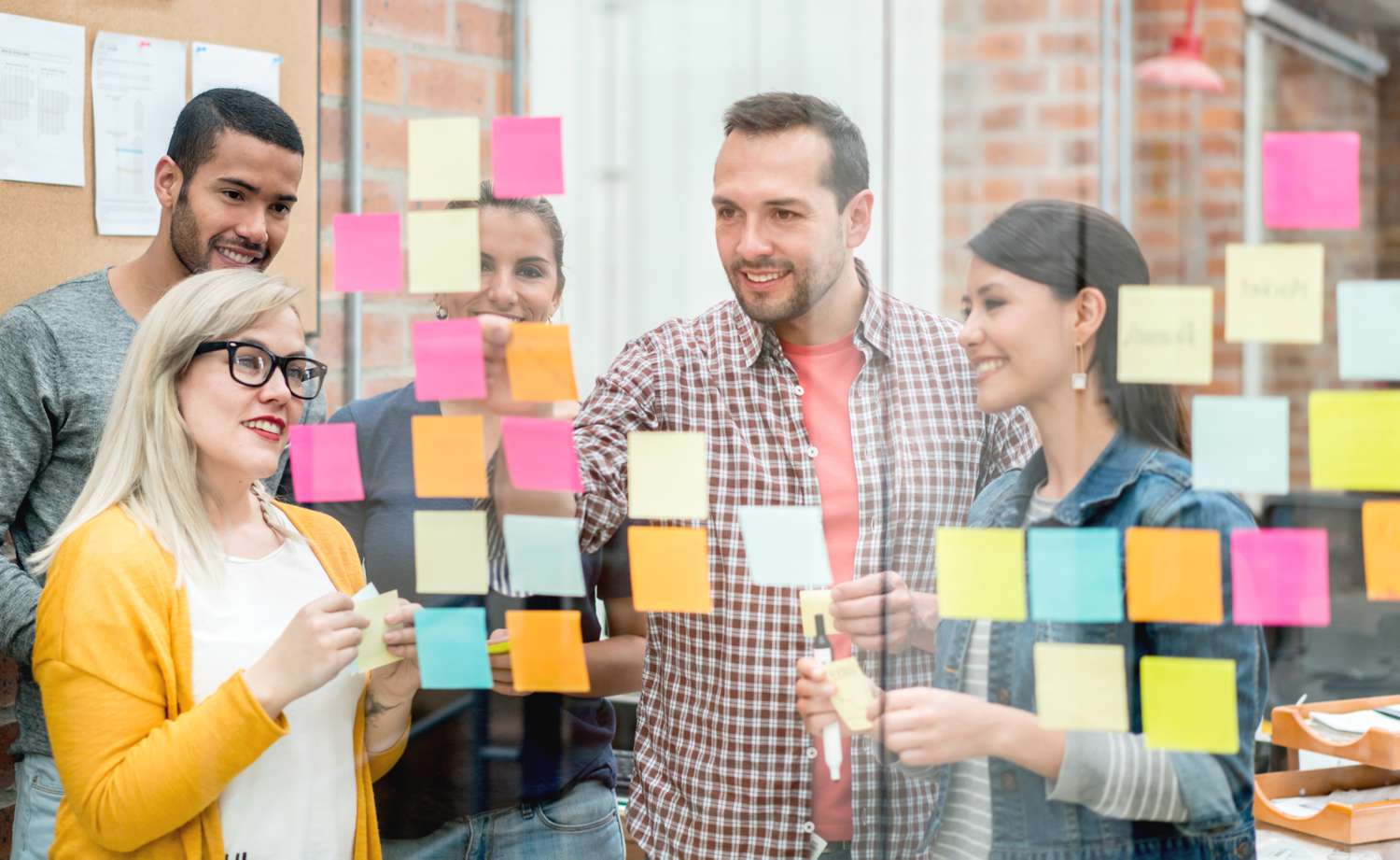 A group of coworkers in an office place sticky notes displaying customer demographics and personality into groups on a large glass room divider.