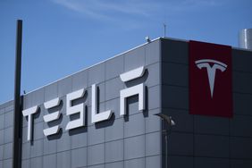 The Tesla logo is seen on the outside of a gray building, with blue sky in the background.