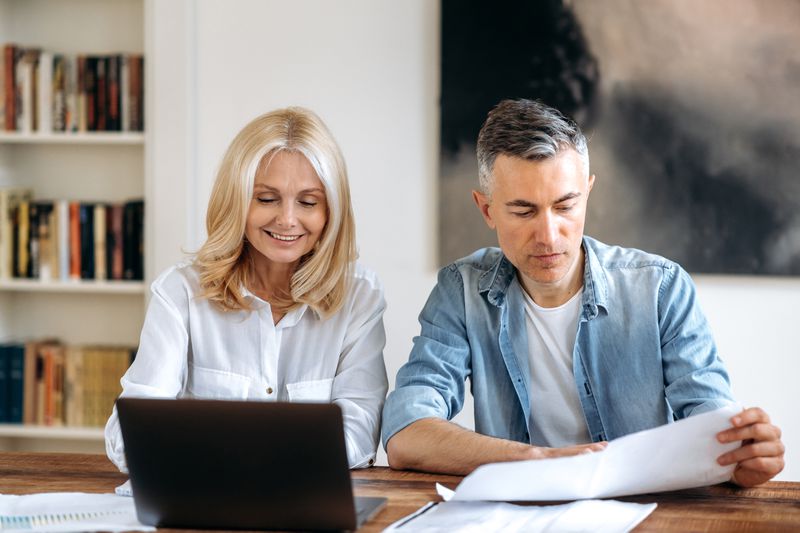 Couple in their 50s sitting at their dining table and looking at mortgage documents and a laptop