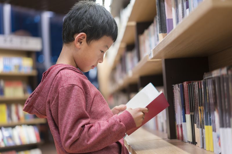 little boy in red hoodie stands up to read book in public library
