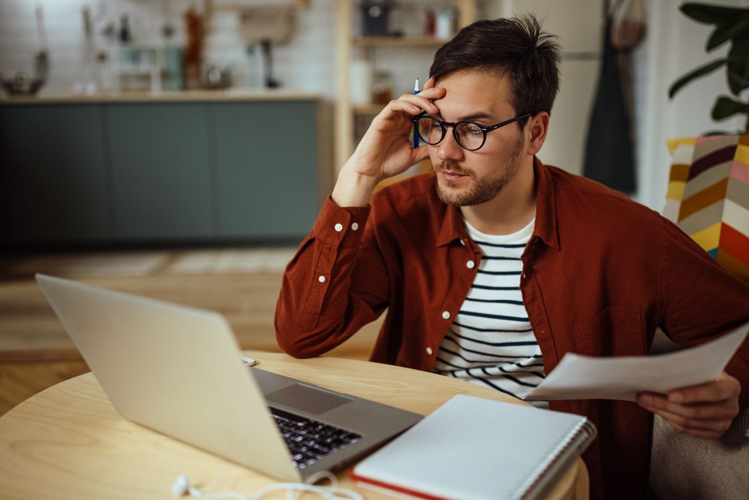 Young man intently studying financial papers and laptop at his kitchen table