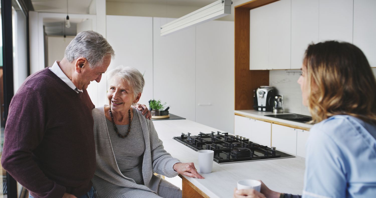 An older man and older woman stand in a kitchen with mugs talking to a younger women