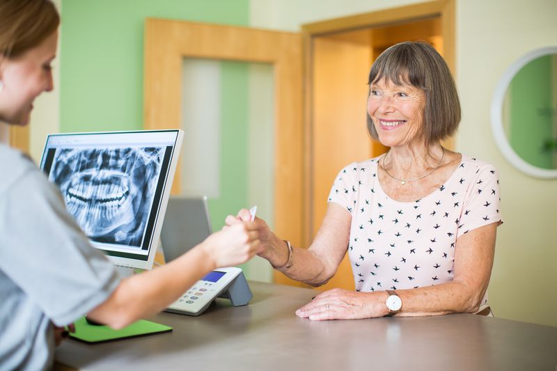 Smiling older woman giving medical card to receptionist who has dental X-rays pulled up on screen
