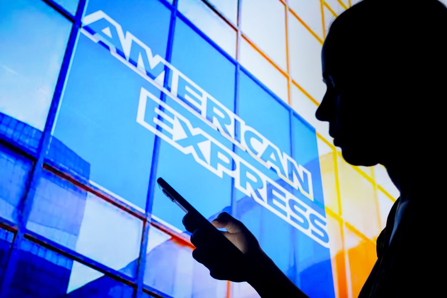  a woman's silhouette holds a smartphone with the American Express (Amex) logo in the background. 