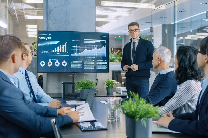 man presenting to a group of people in a conference room with a monitor that shows charts and graphs