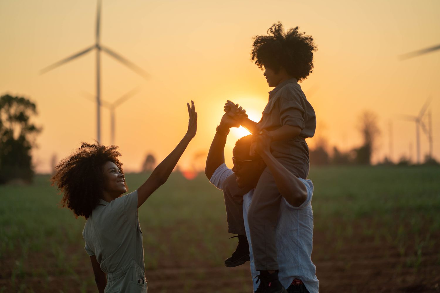  A man holds a child on his shoulders, while another person reaches up to high-five the child, with renewable energy windmills in the background. 