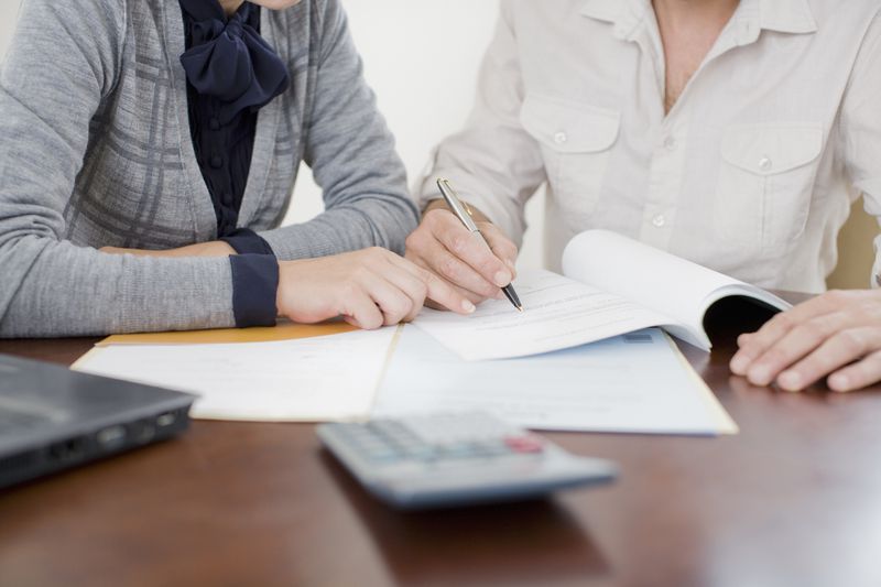 Cropped midsection of two individuals signing papers on a desk with a laptop and calculator on the desk