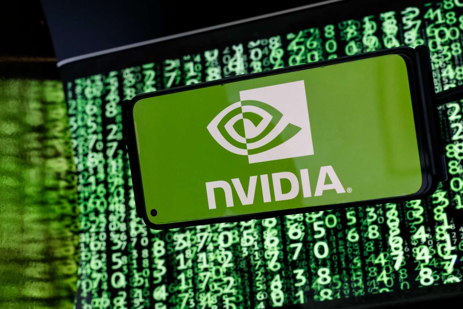 The Nvidia logo is seen displayed on a smartphone screen against a computer screen displaying a matrix image 