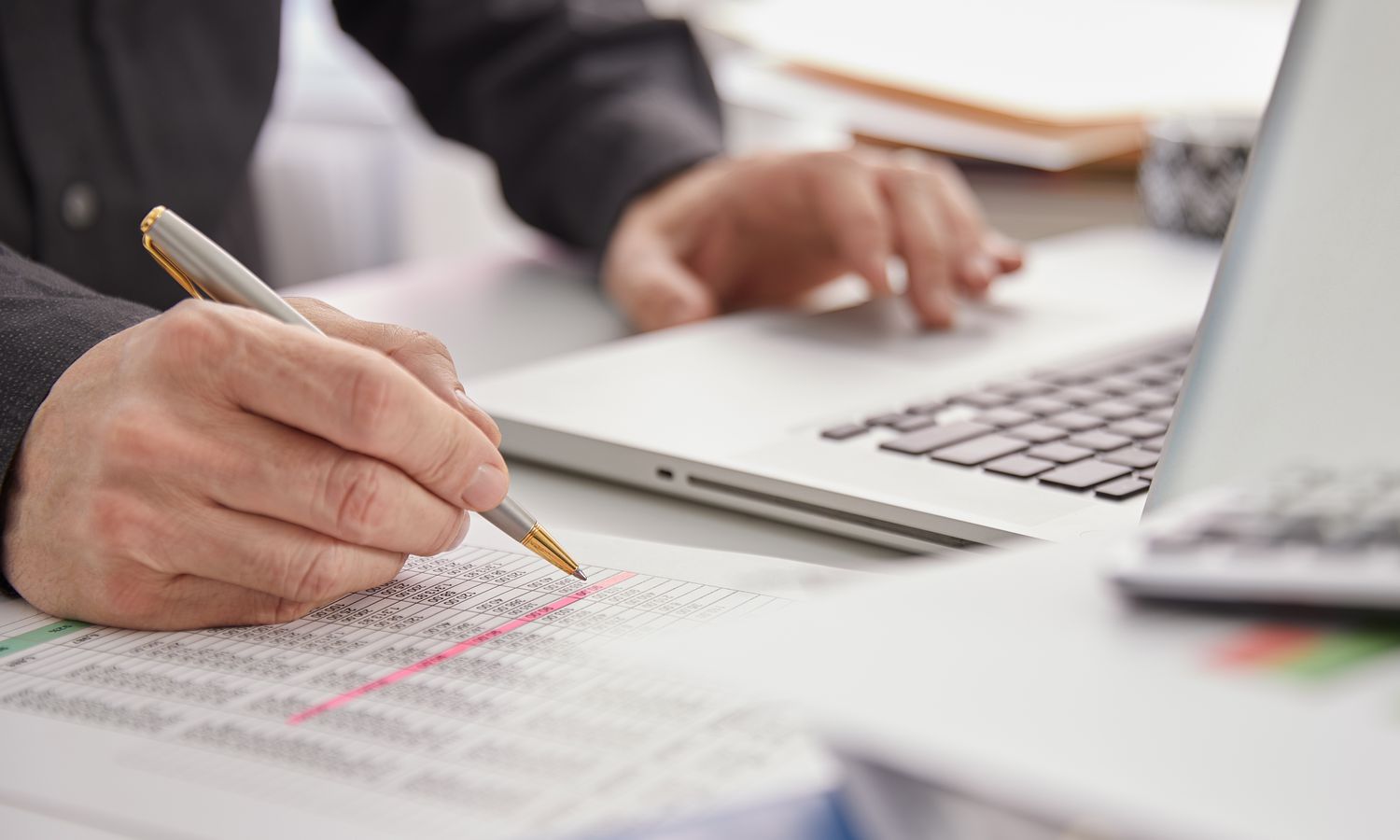 Close up of person reviewing financial sheets with pen with one hand on a laptop computer