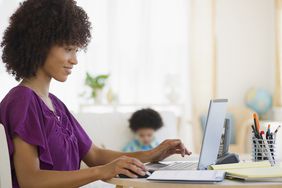 Young mom sitting at a home desk and smiling at something on her laptop, while her young child sits in the background.