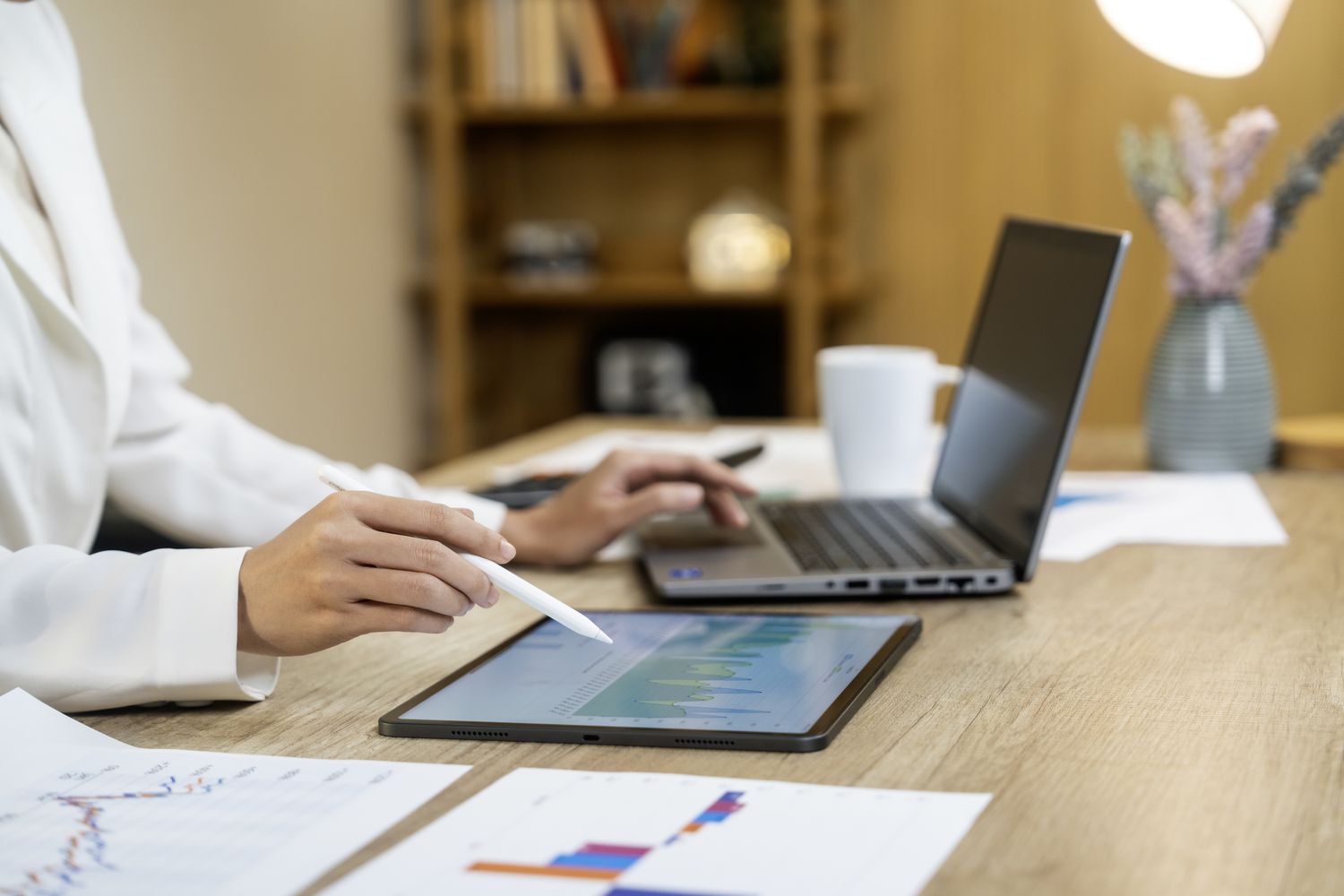 A close-up of a person's hands using a stylus pen to point to graphs on a tablet, while sitting in front of a laptop.