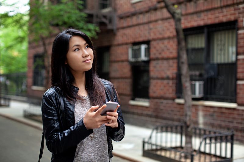 A woman walks on a street outside an apartment building holding her cell phone in her hand