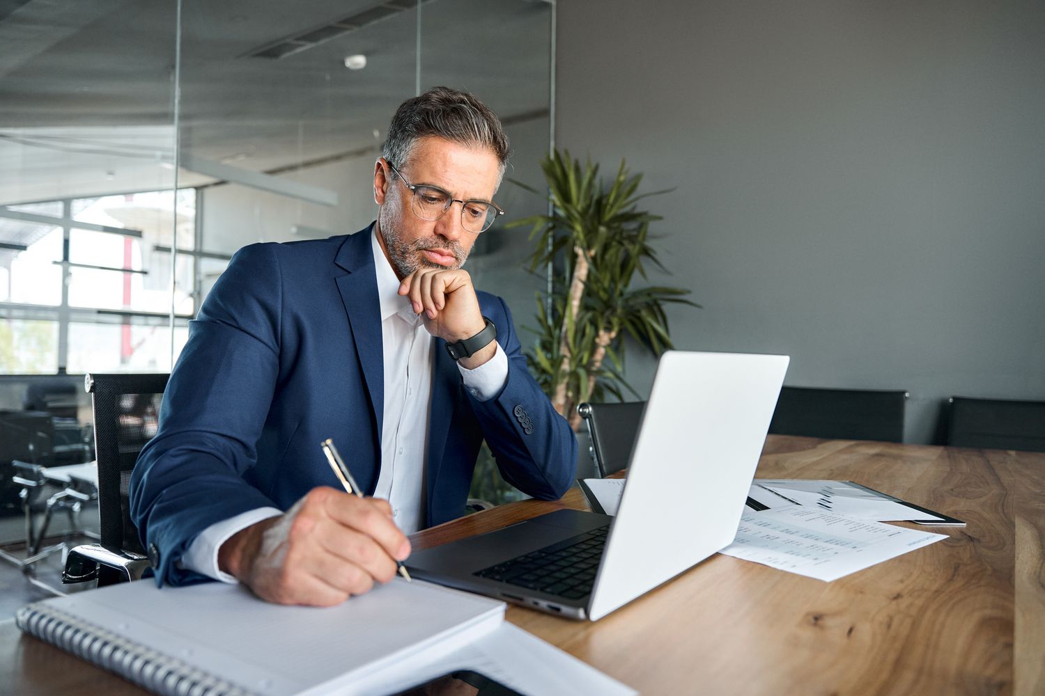 A man in a suit writing in a notebook in front of a laptop in an office. 
