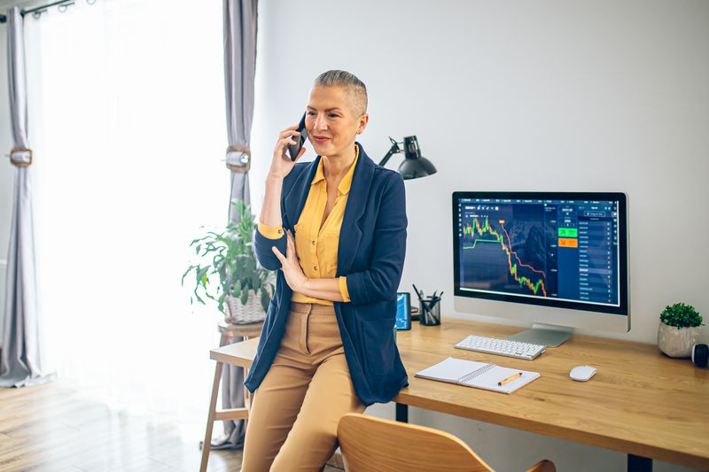 Woman leaning on a desk with her back to a computer monitor, talking on the phone. 