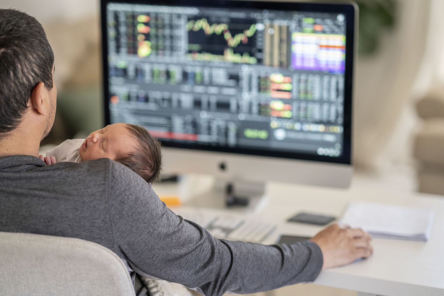 Man looks at market trading screen while holding a newborn baby at home.