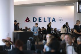 A group of people stand in an airport in front of a Delta Air Lines sign on the wall.