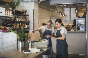 Two business owners wearing aprons look at a tablet and new shipment of goods in their cafe