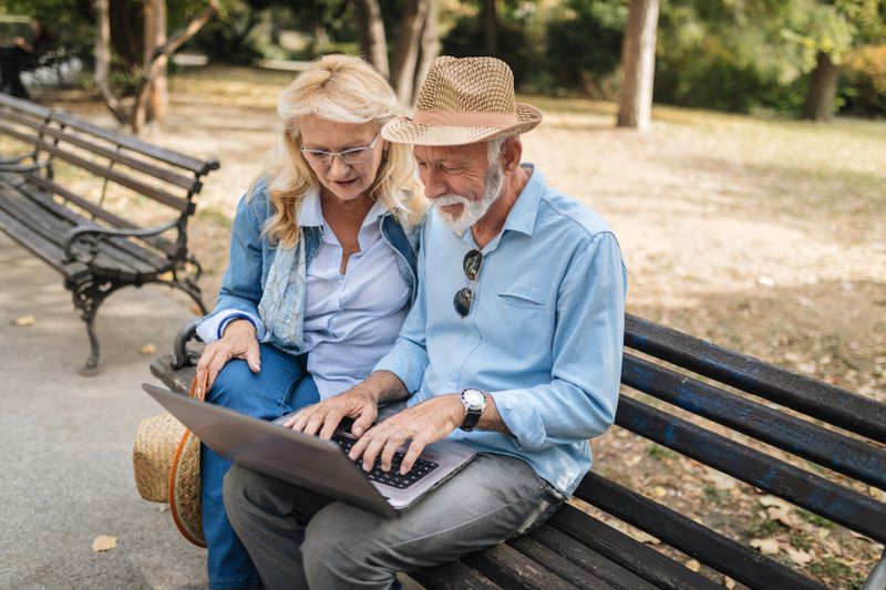 Two annuitants sit on a park bench and use a laptop computer and check the balance of their whole life annuity account.
