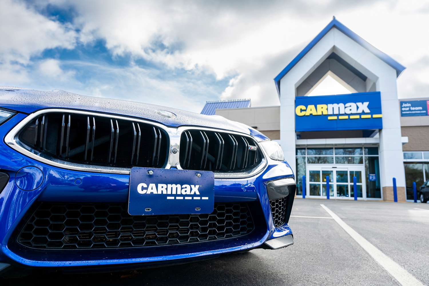 The CarMax logo is seen on a license plate holder on the front of a blue car, with a CarMax dealership in the background.