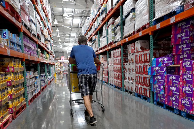 A man with his back to the camera pushes a shopping cart down an aisle at Costco.
